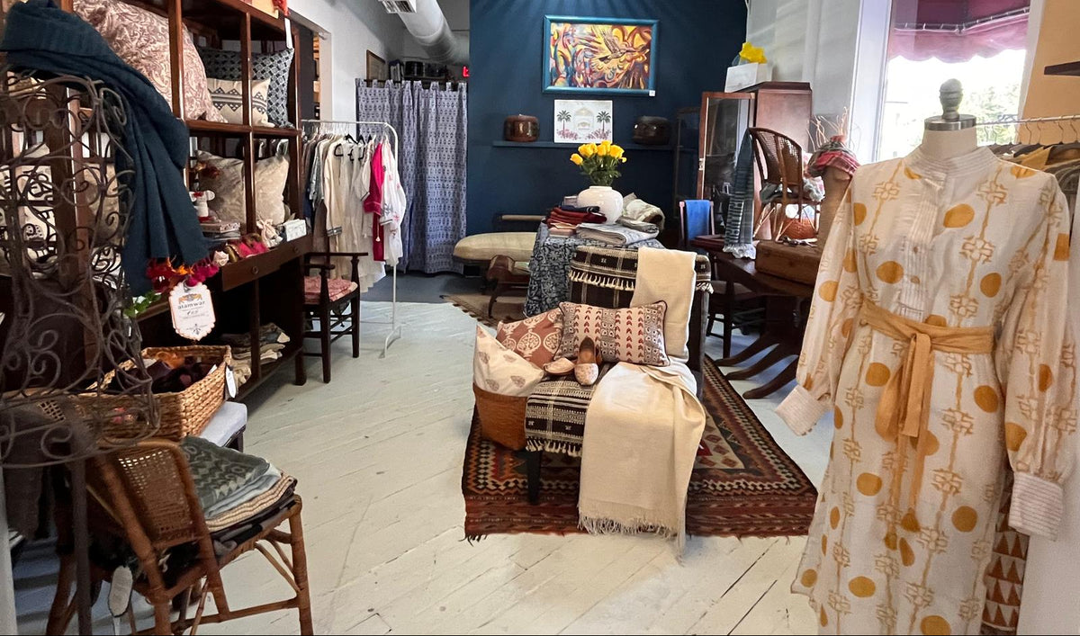 A boutique interior with multiple home decor and textile items displayed throughout. A cream robe with a mustard yellow circular pattern is shown on a mannequin in the foreground right, while shelves, chairs, pillows, and a patterned wall display are visible in the room.