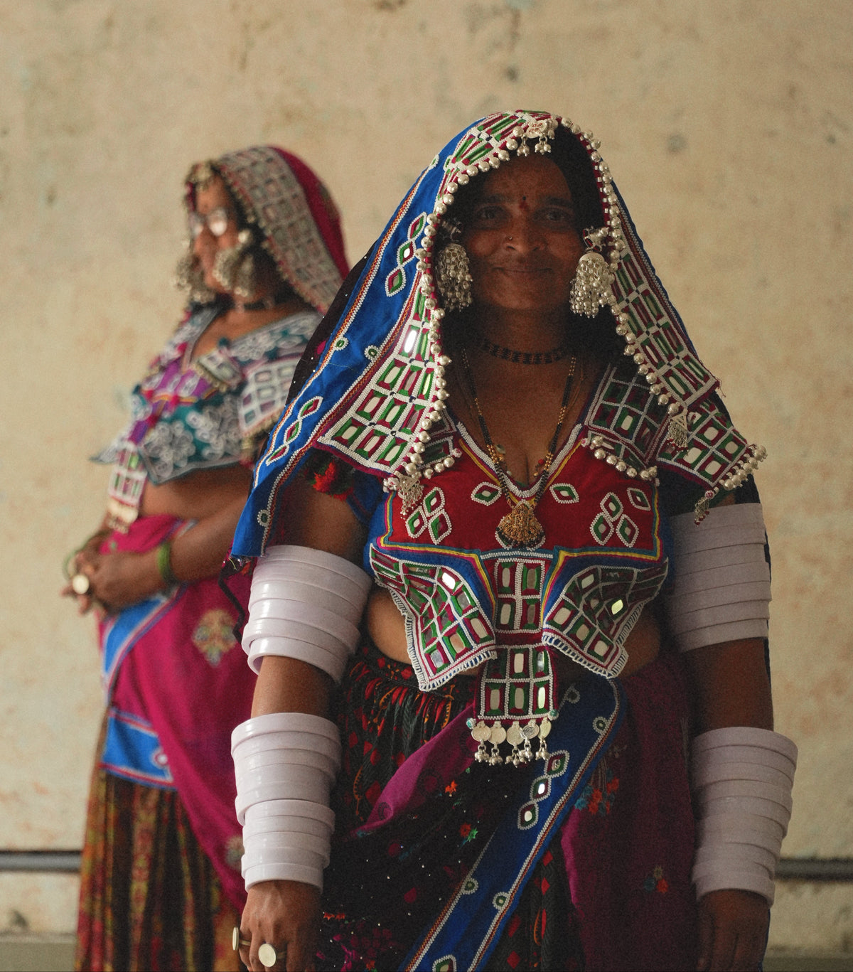 A woman in traditional Indian attire stands in front of a textured beige wall, wearing a colorful embroidered sari with a blue and red patterned drape, silver mirror work, and white arm wraps. Another woman is softly blurred in the background.