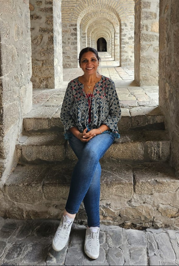 Chitra Reddy wearing a patterned long-sleeve blouse, blue jeans, and white sneakers sits on stone steps inside a historic stone structure with repeating brick arches.