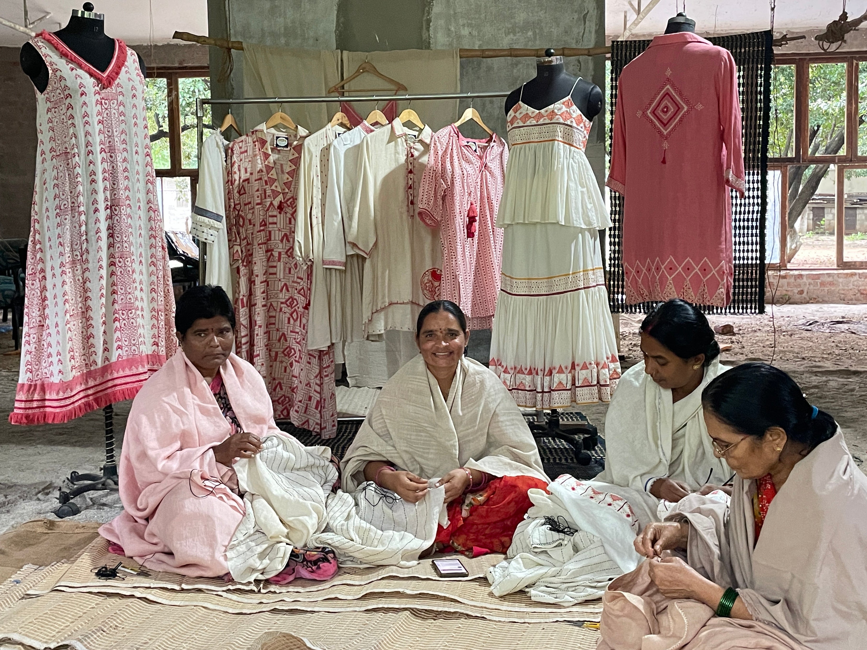 Four women sitting on the floor surrounded by fabric and clothing in a workshop setting.