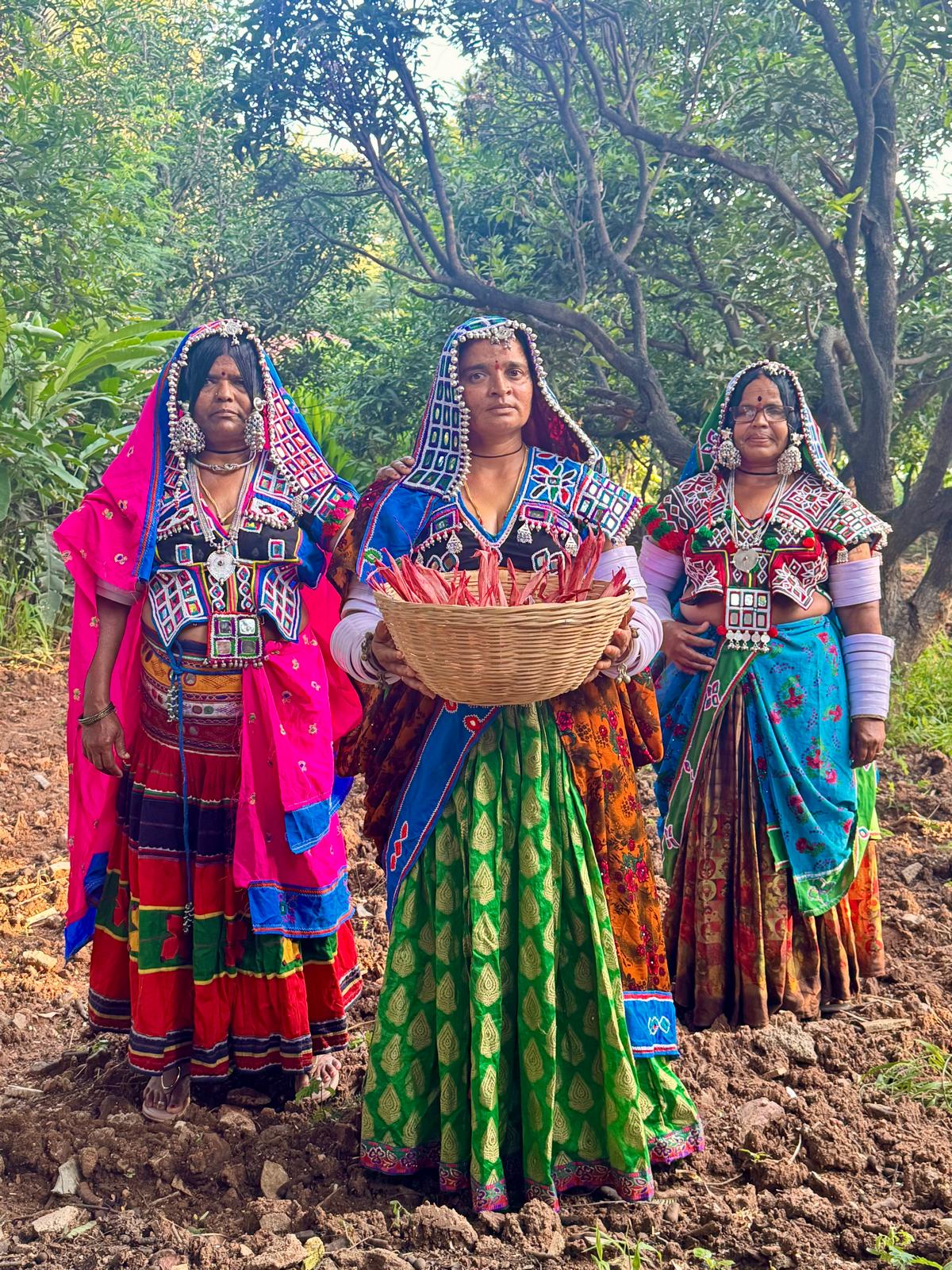 Three women in colorful traditional ethnic clothing stand in a muddy outdoor setting with trees behind them. The central woman holds a wicker basket filled with red chili peppers.