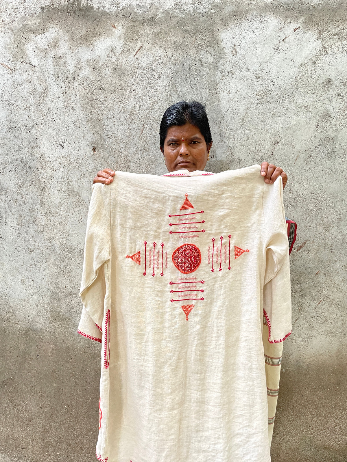 A person holds up a cream-colored cotton garment with red geometric embroidery against a textured concrete wall.
