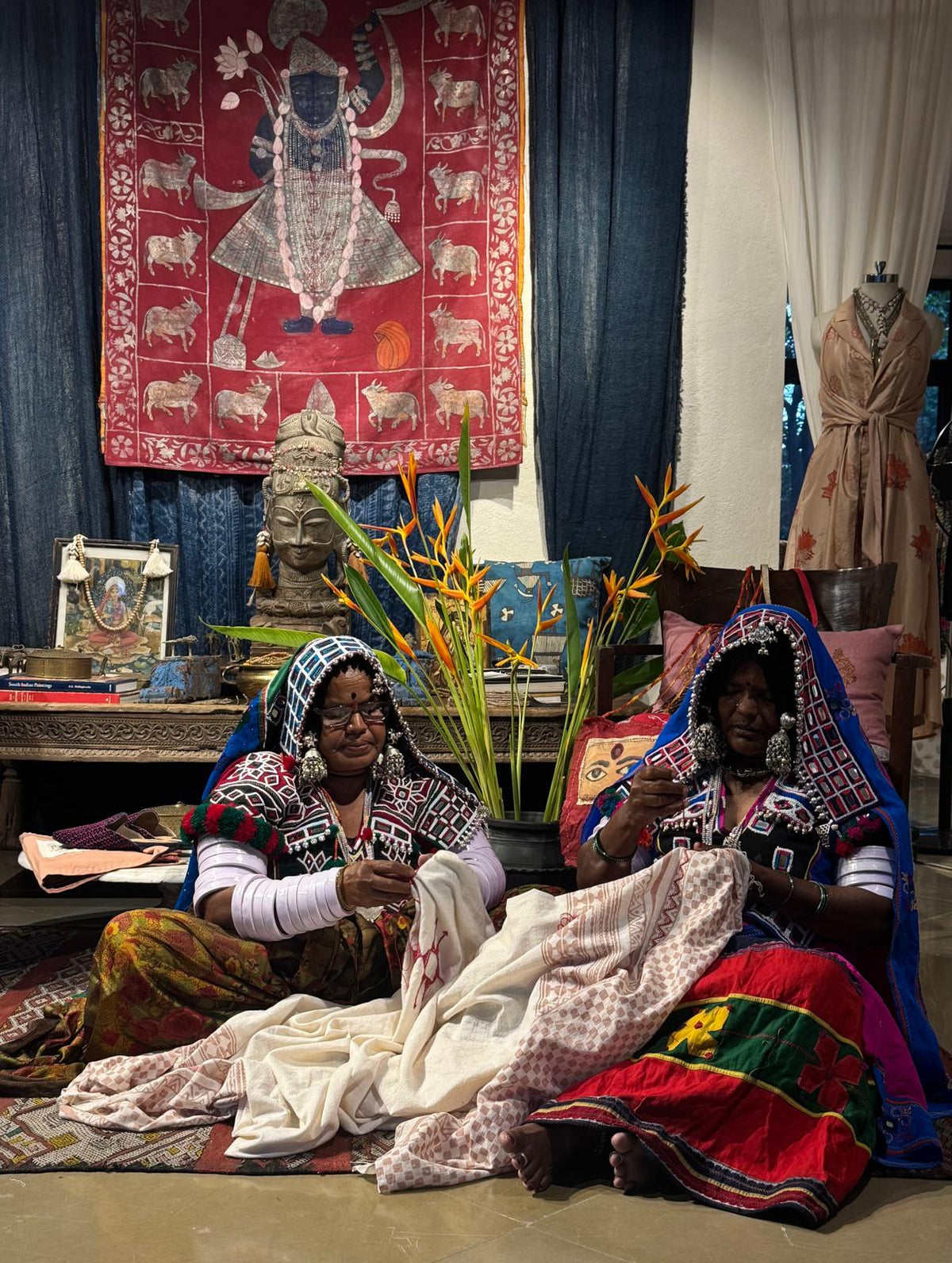Two women seated indoors on a patterned rug, handling a cream-colored patterned textile. The scene is decorated with colorful traditional garments, a large red tapestry, blue curtains, and a potted plant with orange flowers in the center.