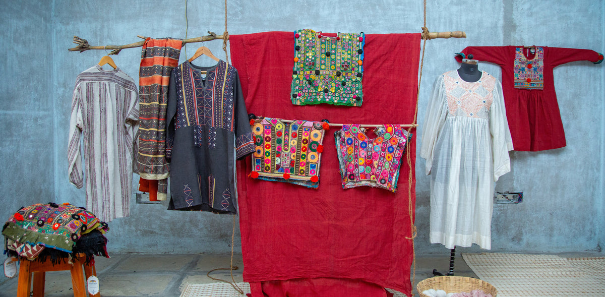A collection of traditional embroidered garments and textiles displayed in a rustic room with a blue-gray concrete wall. A large red fabric backdrop hangs in the center, flanked by hanging garments including a white embroidered dress, a red embroidered tunic, a striped shirt, and patterned scarves, with additional folded textiles on a wooden stool.