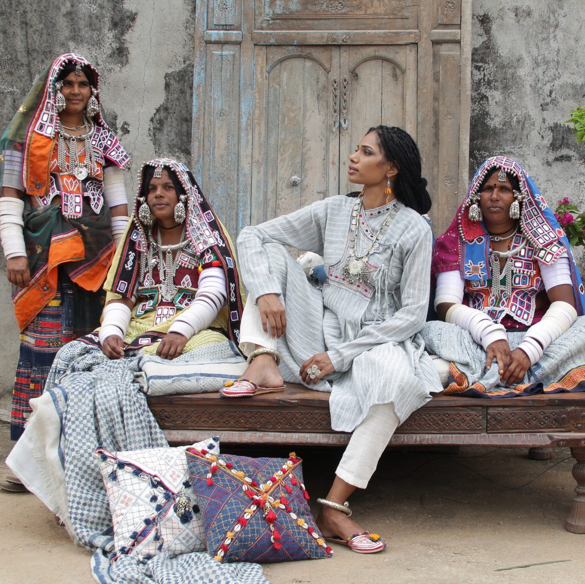 Four women in traditional South Asian attire sit and stand on a carved wooden bench in front of a weathered wooden door. They wear ornate jewelry, colorful embroidered headscarves, and patterned garments, with decorative pillows on the ground in the foreground.