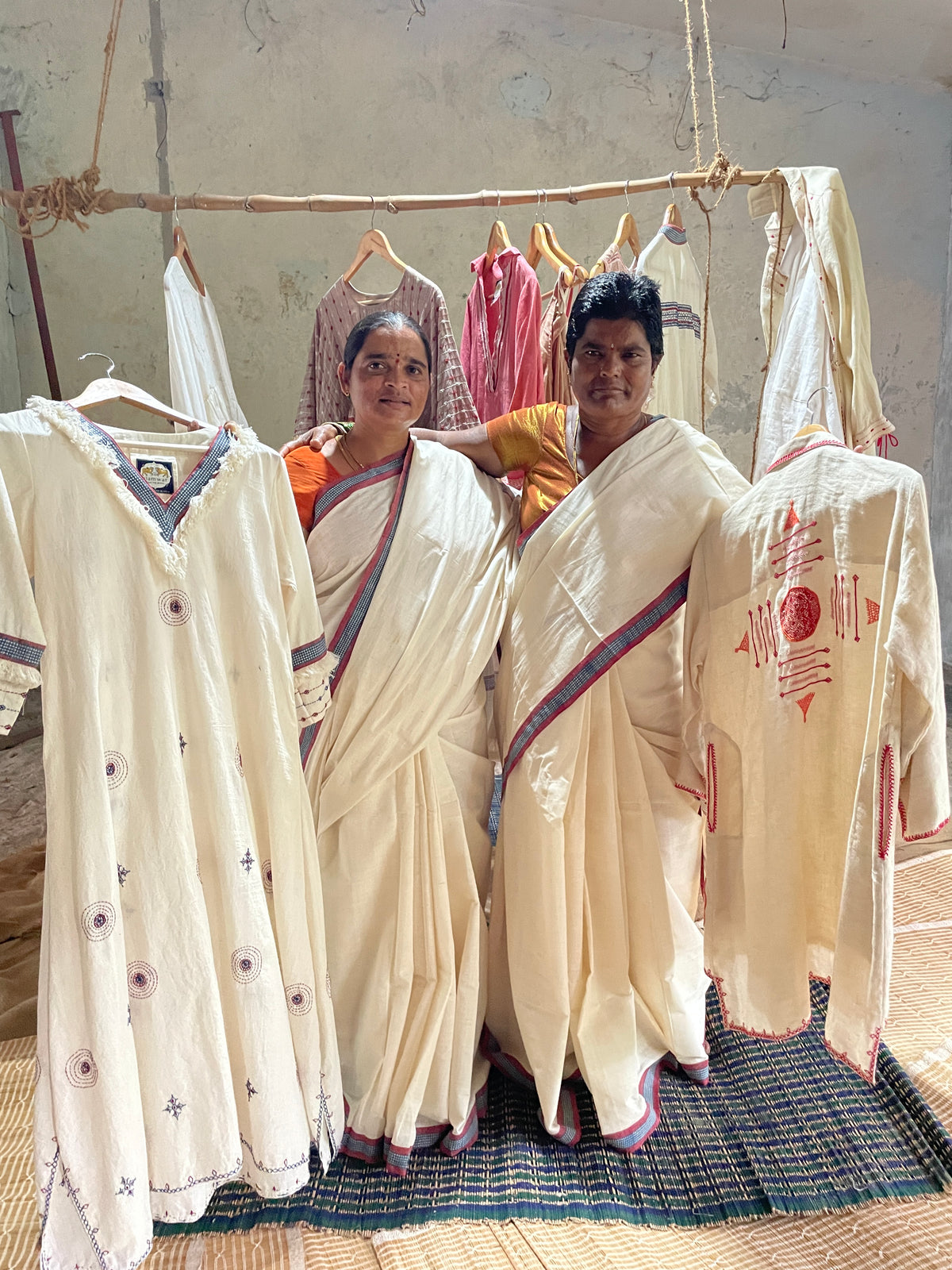 Two women wearing cream-colored sarees stand behind hanging cream-colored embroidered garments in a rustic indoor setting. The foreground shows detailed close-ups of the embroidered clothing, including a cream kurta with purple floral motifs and a cream saree with a blue and red border.