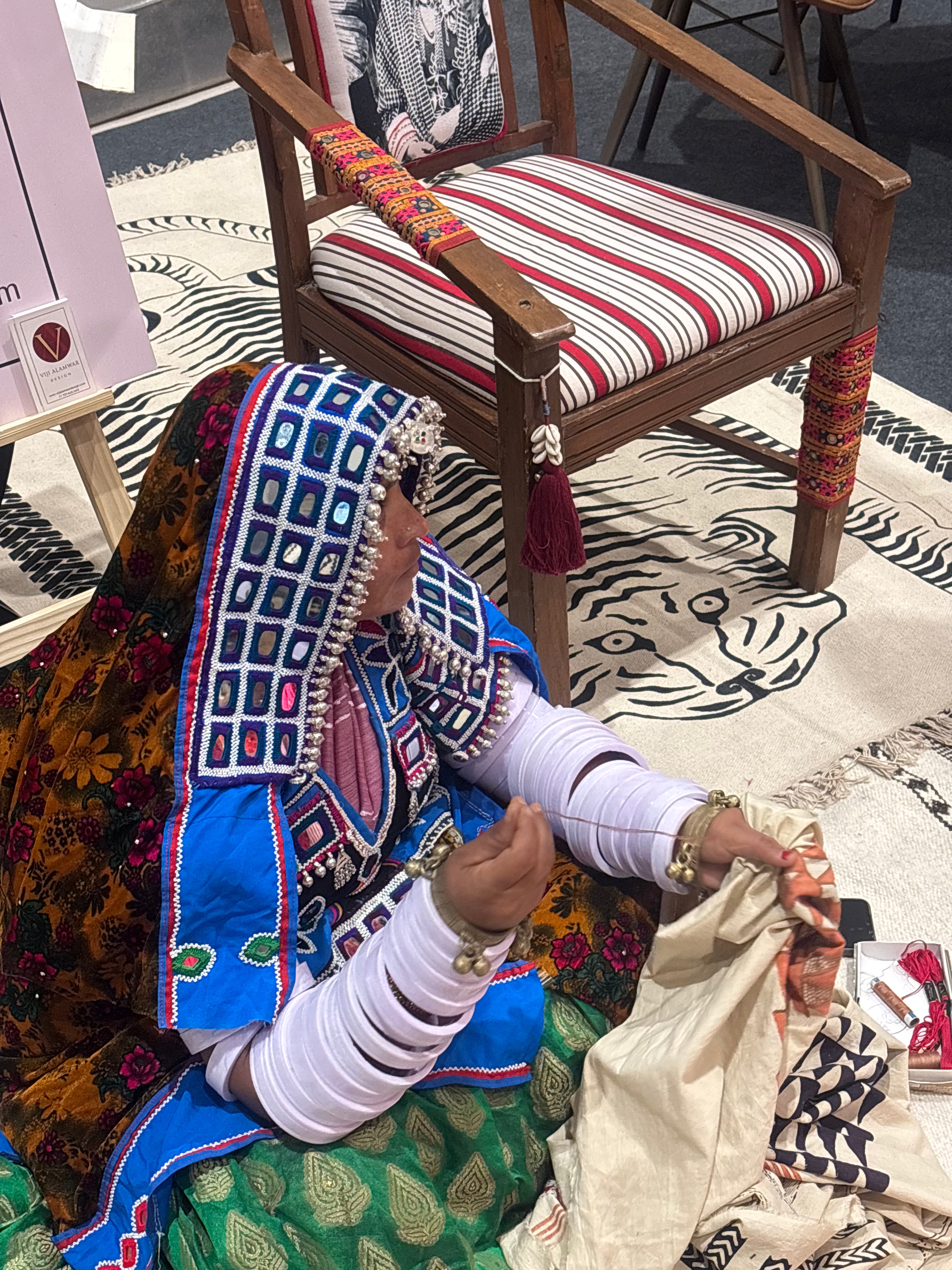 A woman in traditional South Asian attire with intricate blue, red, and white embroidery sits on a patterned rug next to a wooden armchair with a striped cushion.
