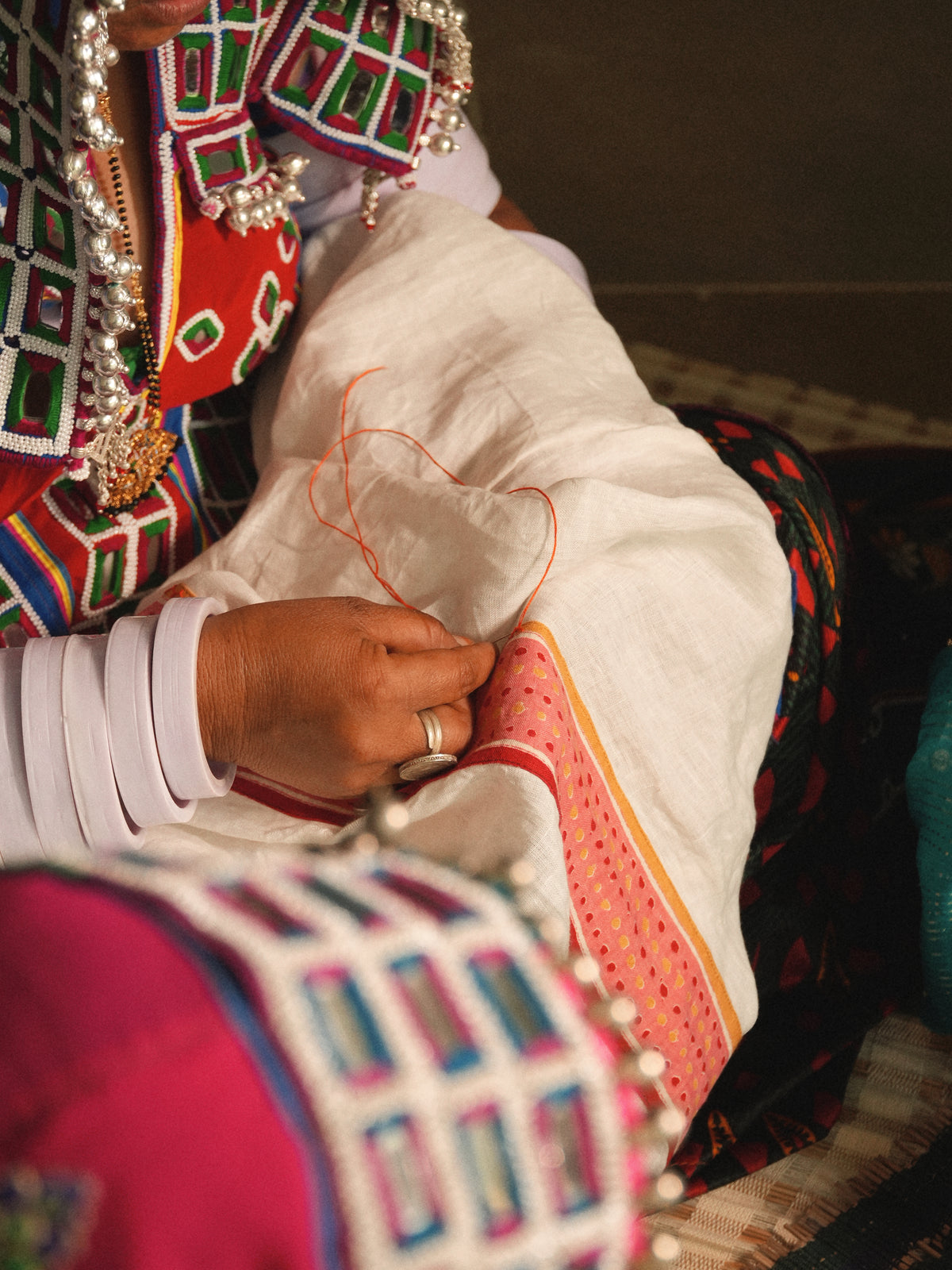 A close-up of a person's hands sewing a white garment with orange thread, wearing traditional colorful embroidered clothing and silver jewelry.
