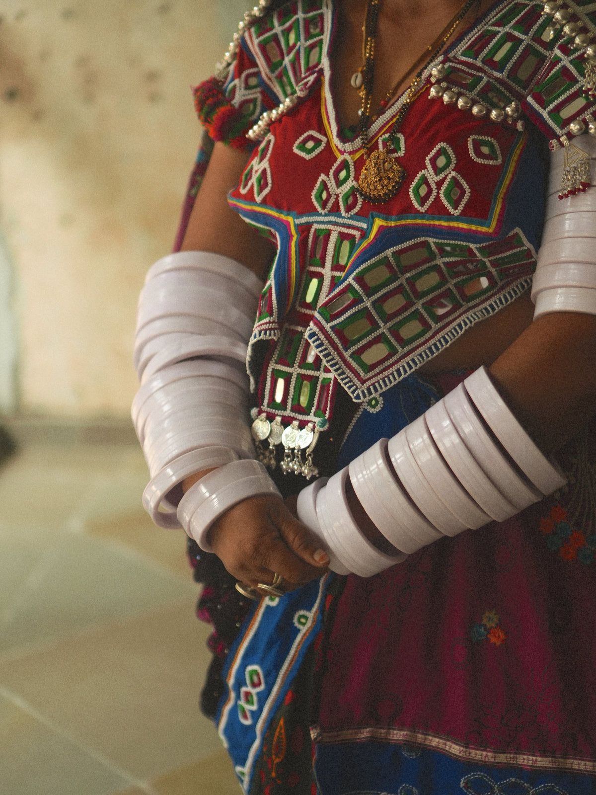 A close-up of a banjara person wearing a colorful embroidered traditional outfit with layered white bangles on both forearms and gold jewelry, standing indoors against a softly blurred neutral background.